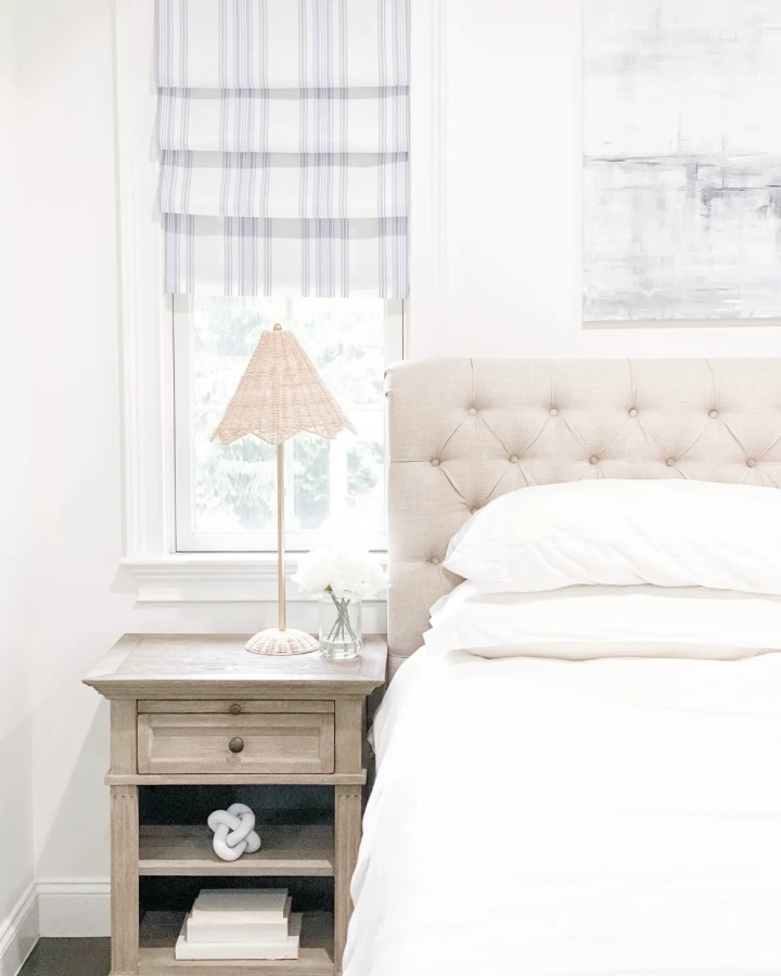 partial view of a bedroom with a beige upholstered headboard, white bedding, a beige night stand and a striped faux roman shade hanging in the window above it