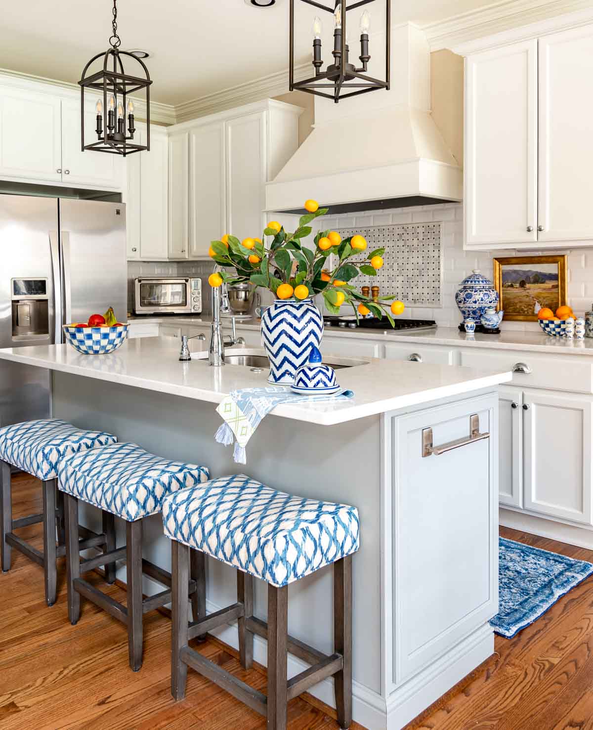gray island in a kitchen with white cabinets. bar height stools upholstered in blue and white fabric. blue and white accents on the counters and a vase filled with faux orange stems on the island