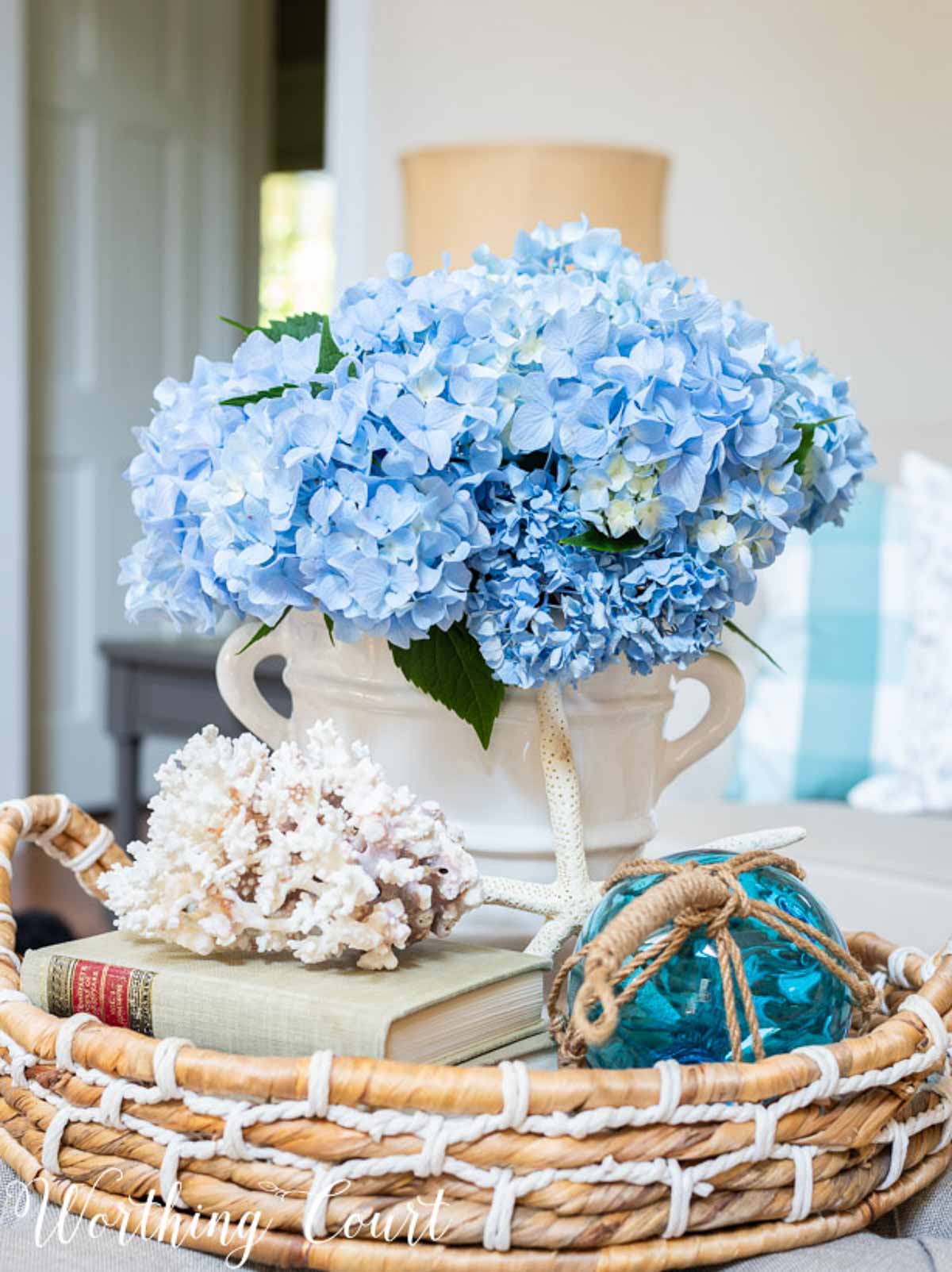 bunch of blue hydrangeas arranged in a white vase and displayed in a round wicker basket with a blue float and piece of white coral on top of a book