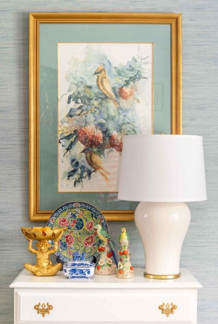 white lamp and chinoiserie accessories on a white chest, underneath traditional style artwork against a wall with blue grasscloth wallpaper
