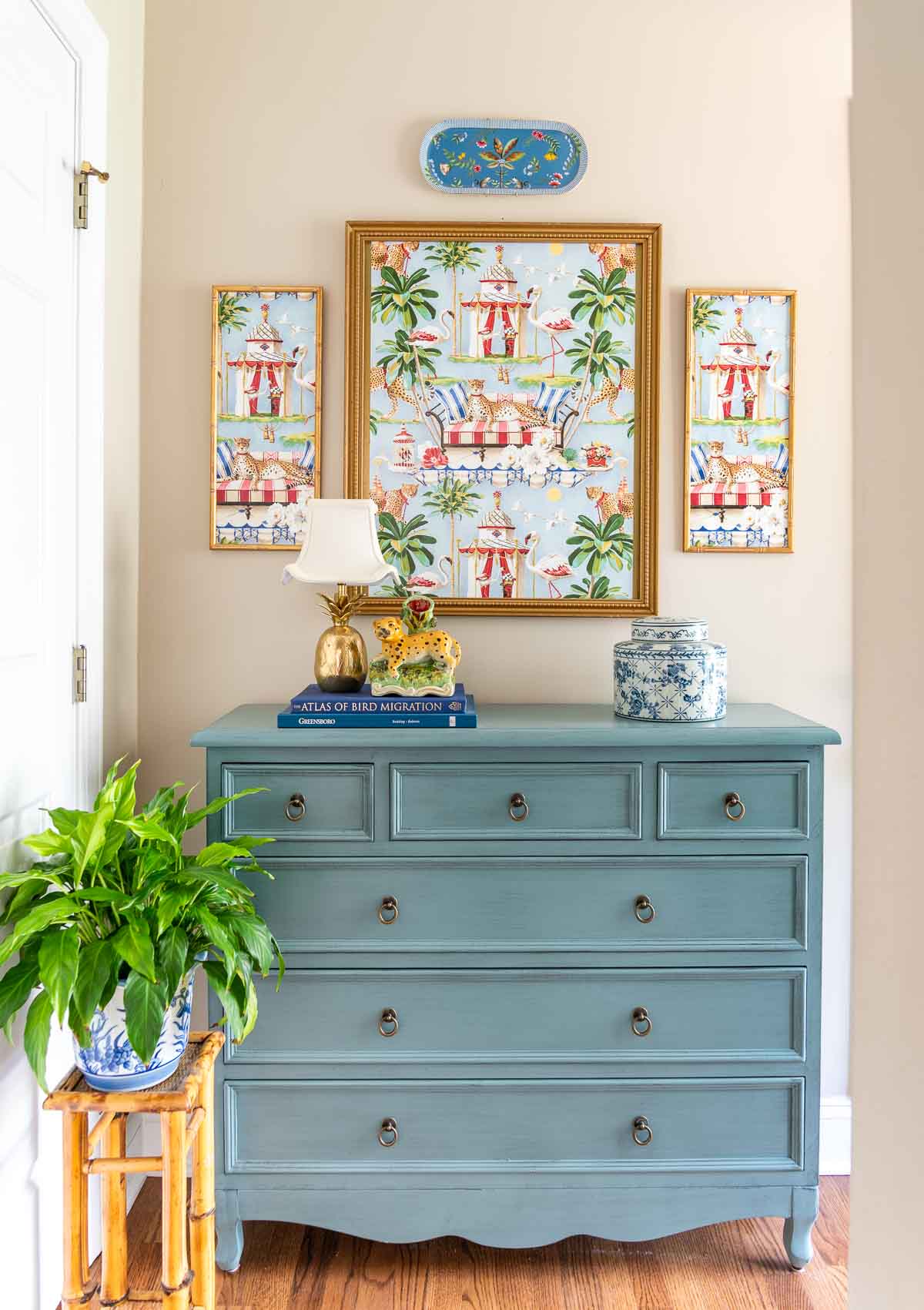 small blue chest in a hallway with a trio of chinoiserie framed art above and chinoiserie type accessories and lamp decorating the top
