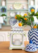 blue and white pagoda shaped lantern and a blue and white vase with faux orange stems on a kitchen table with a decorated plate rack in the background