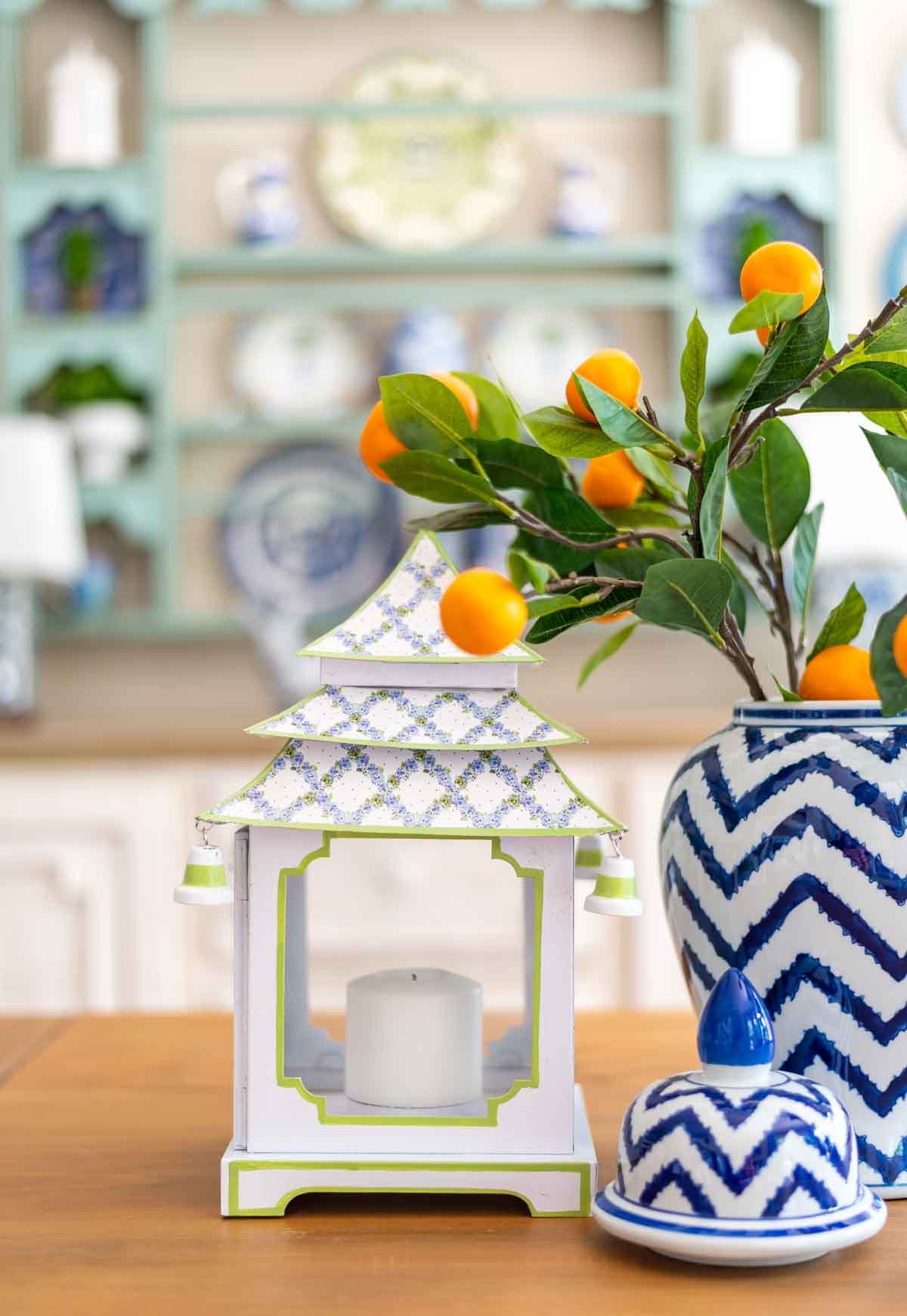 blue and white pagoda shaped lantern and a blue and white vase with faux orange stems on a kitchen table with a decorated plate rack in the background