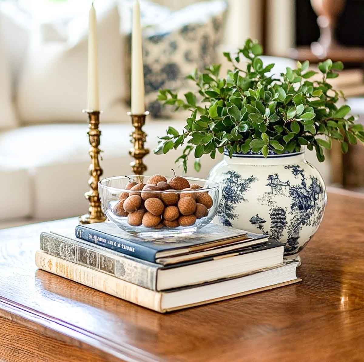 late summer coffee table vignette with a green plant in a blue and white vase, a pair of brass candlesticks and a glass bowl filled with seed pods on top of a stack of books