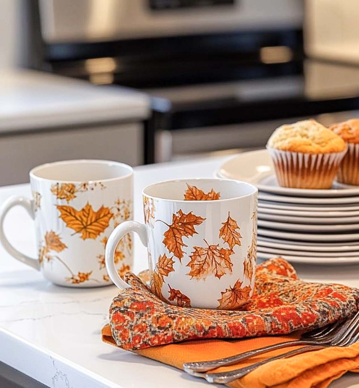 two coffee mugs with fall leaves on them in vignette with fall colored linens on a kitchen counter