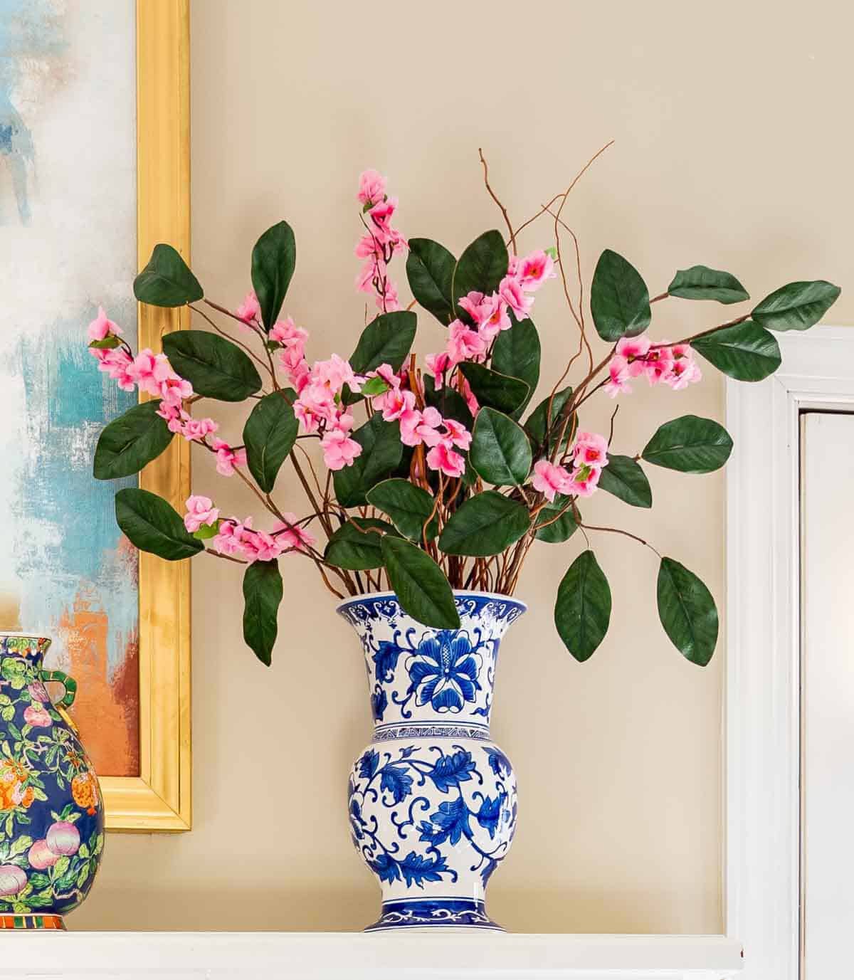 pink faux flower and greenery stems in a tall blue and white vase on a mantel for spring