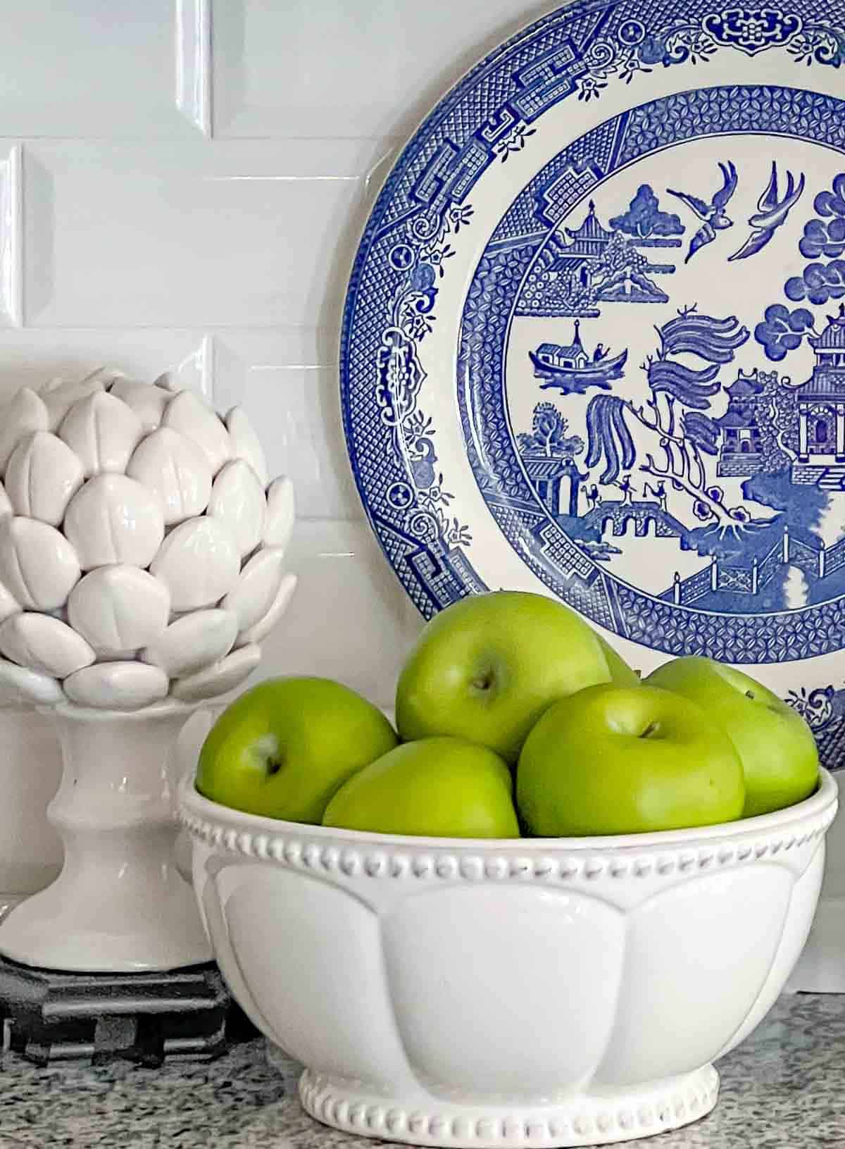 green apples in a white bowl and white artichoke on a kitchen counter in front of a blue and white plate hanging on a white backsplash for spring