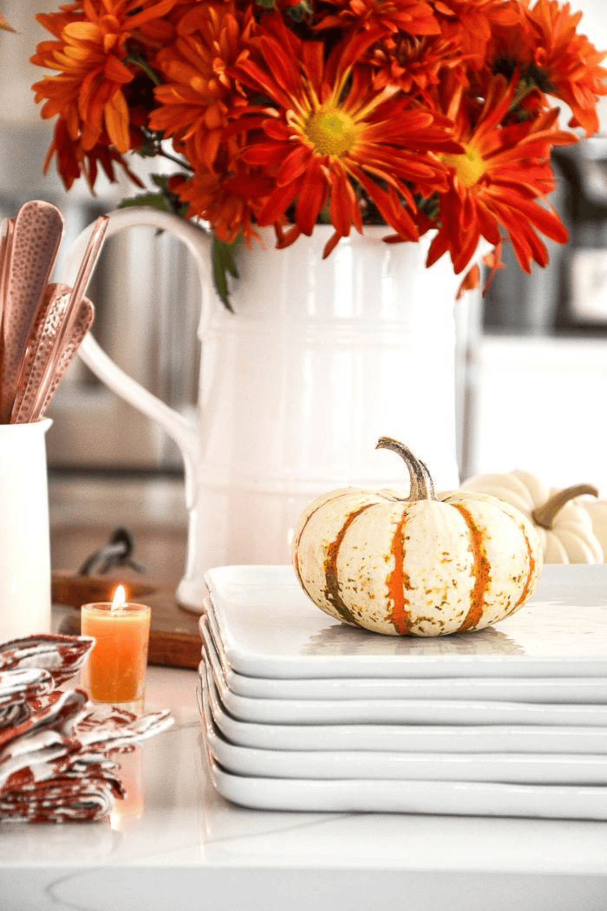 white pitcher filled with fall flowers behind a stack of white dishes with a small pumpkin on top