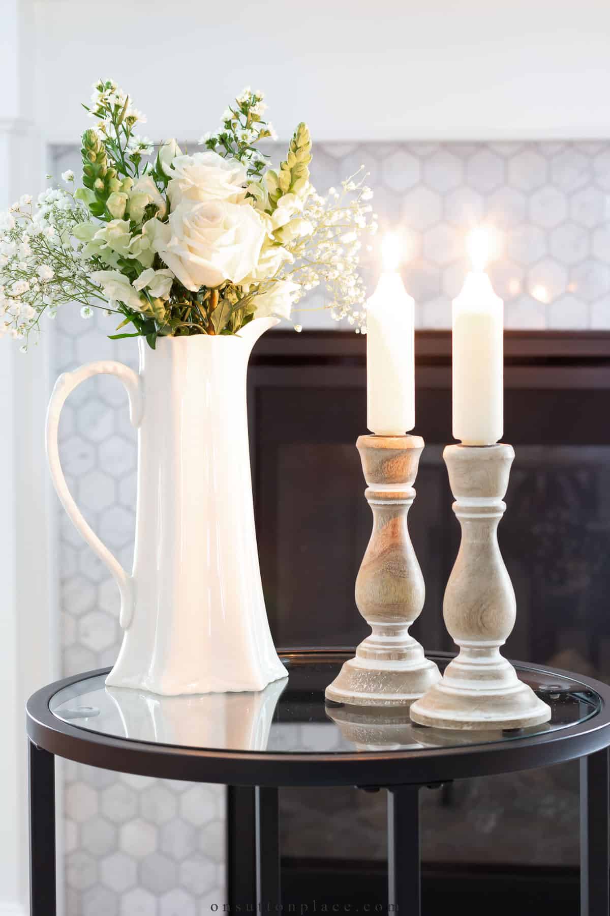 gray candlesticks with white candles and a white pitcher with white flowers in it