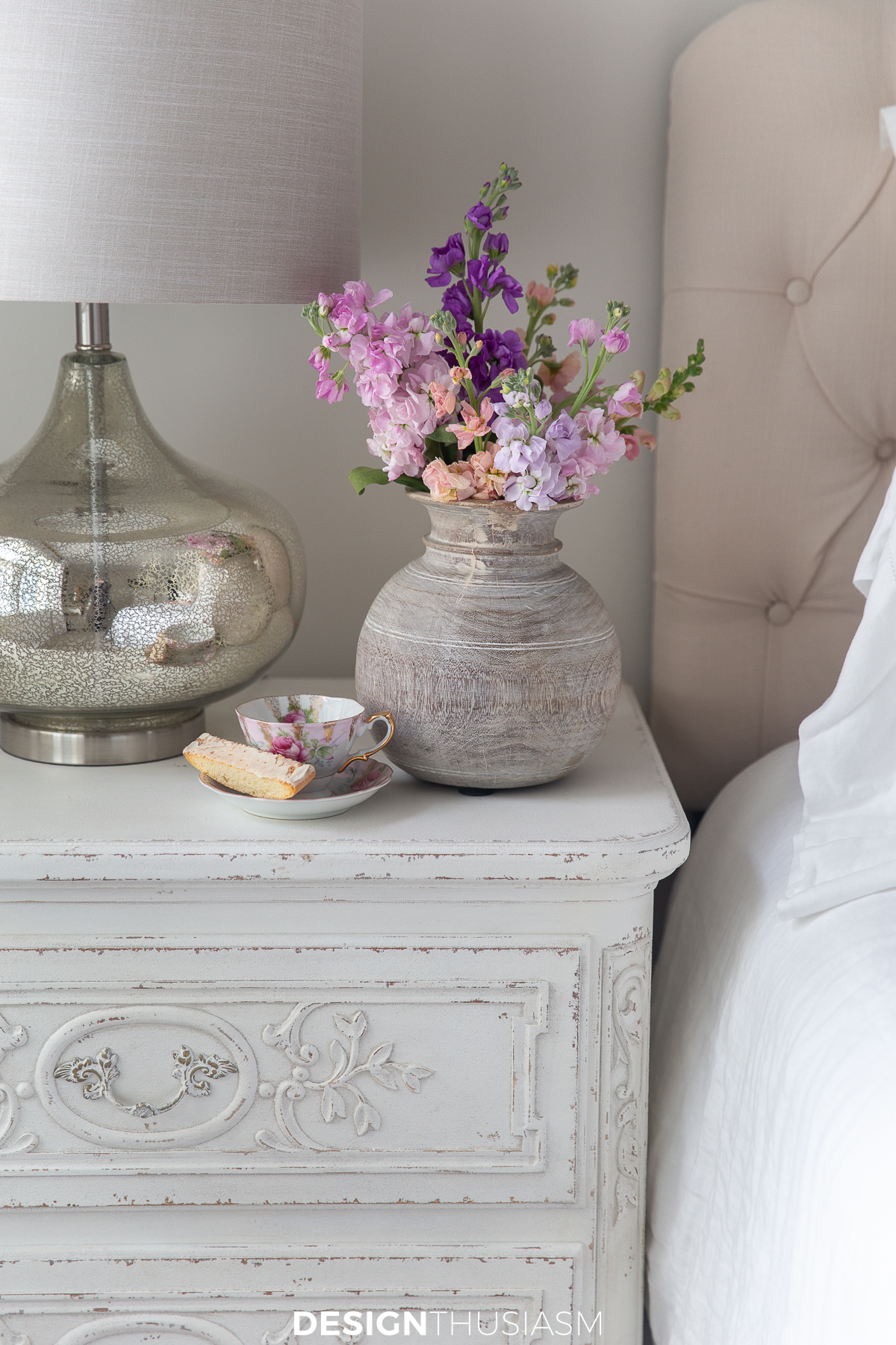 corner of a white nightstand beside a bed with a vase of pink flowers