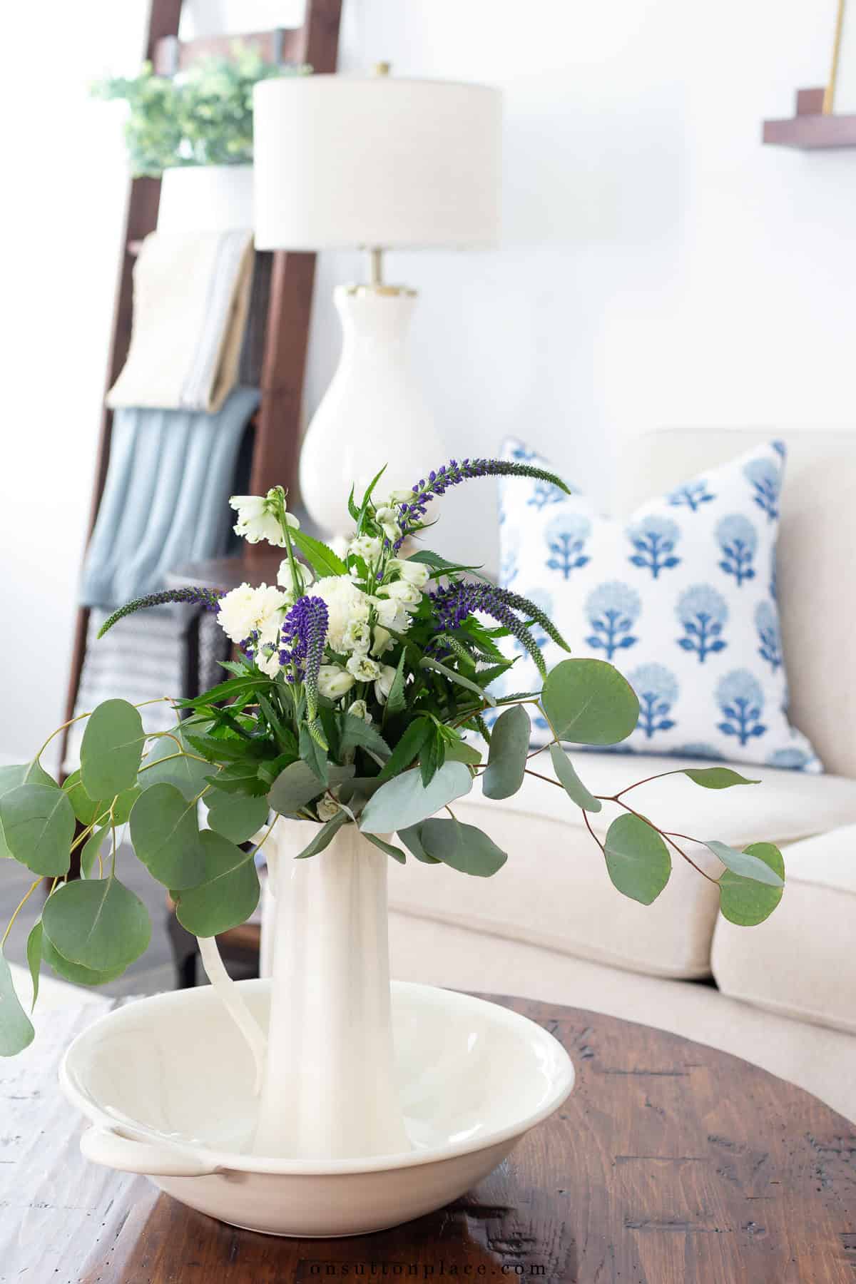 greenery in white vessels on a wood coffee table in front of a white couch with a blue and white throw pillow