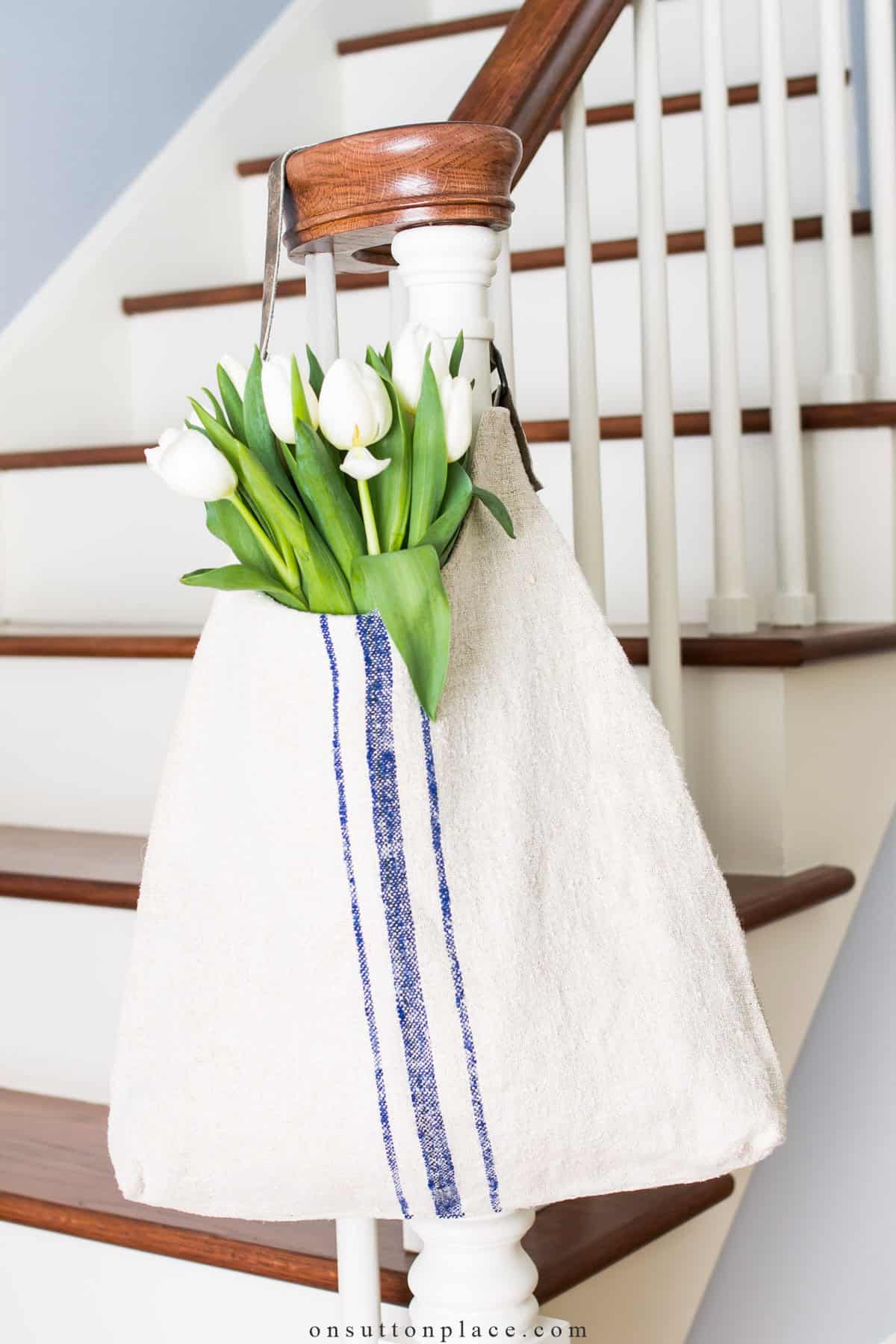 White bag tote back made with grain sack fabric hanging on a newel post with white tulips inside