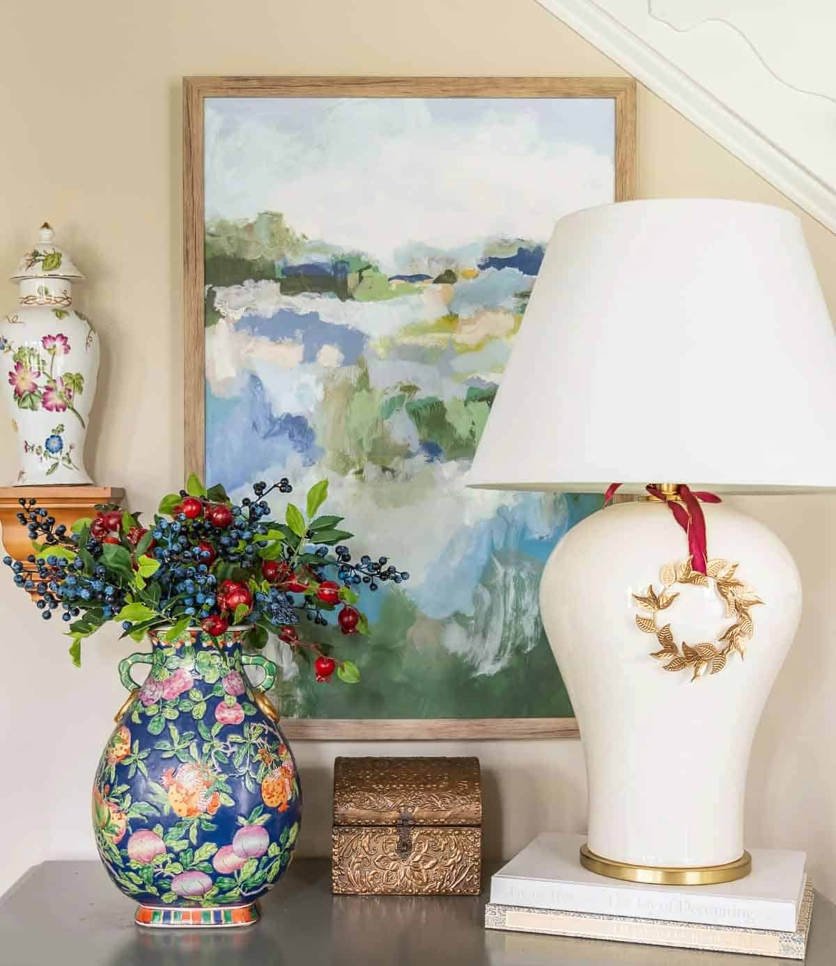 gray chest with a white lamp, blue vase filled with berry stems and abstract wall art above