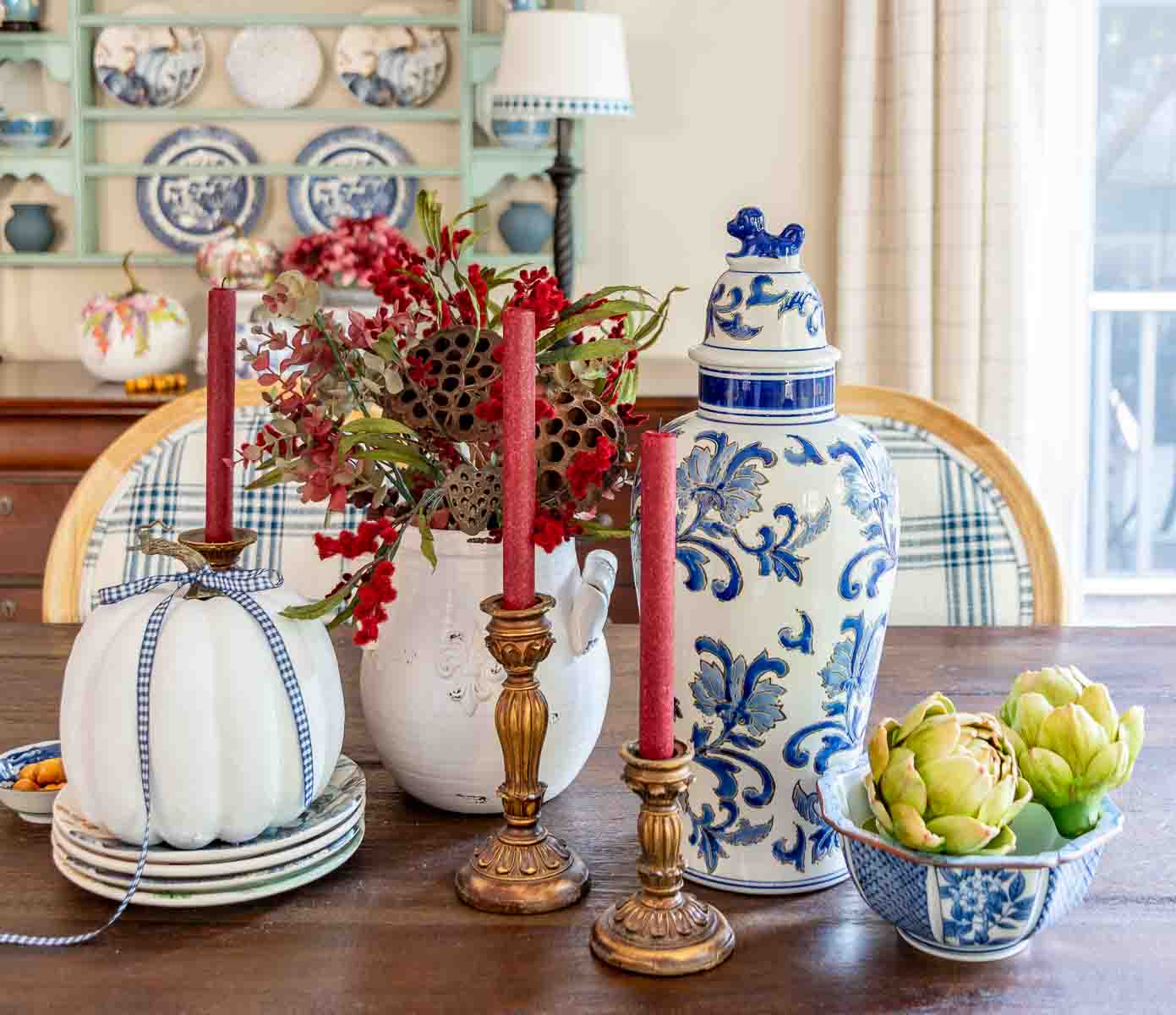 a diy fall table centerpiece using blue, white and burgundy colors, consisting of a large lidded urn, a floral arrangement, and a large white pumpkin on a stack of fall plates