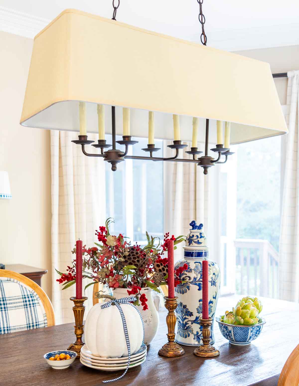 a diy fall table centerpiece using blue, white and burgundy colors, consisting of a large lidded urn, a floral arrangement, and a large white pumpkin on a stack of fall plates