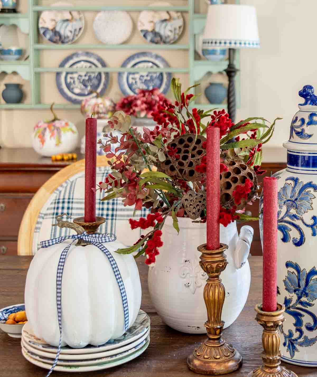 a diy fall table centerpiece using blue, white and burgundy colors, consisting of a large lidded urn, a floral arrangement, and a large white pumpkin on a stack of fall plates