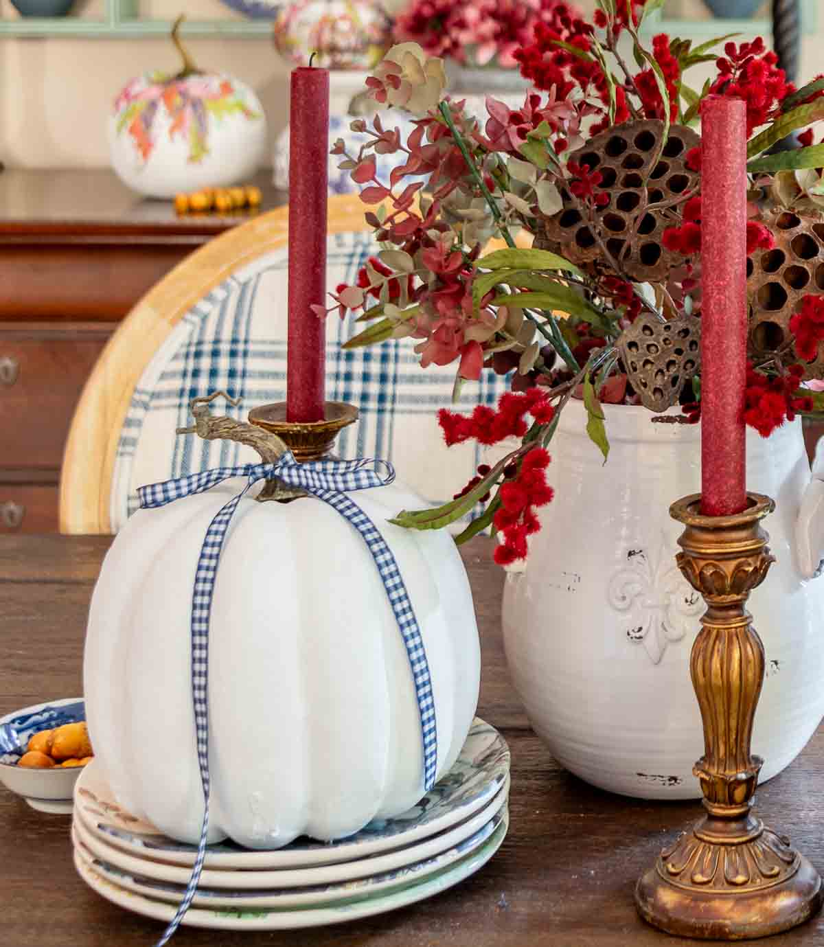 a diy fall table centerpiece using blue, white and burgundy colors, consisting of a large lidded urn, a floral arrangement, and a large white pumpkin on a stack of fall plates