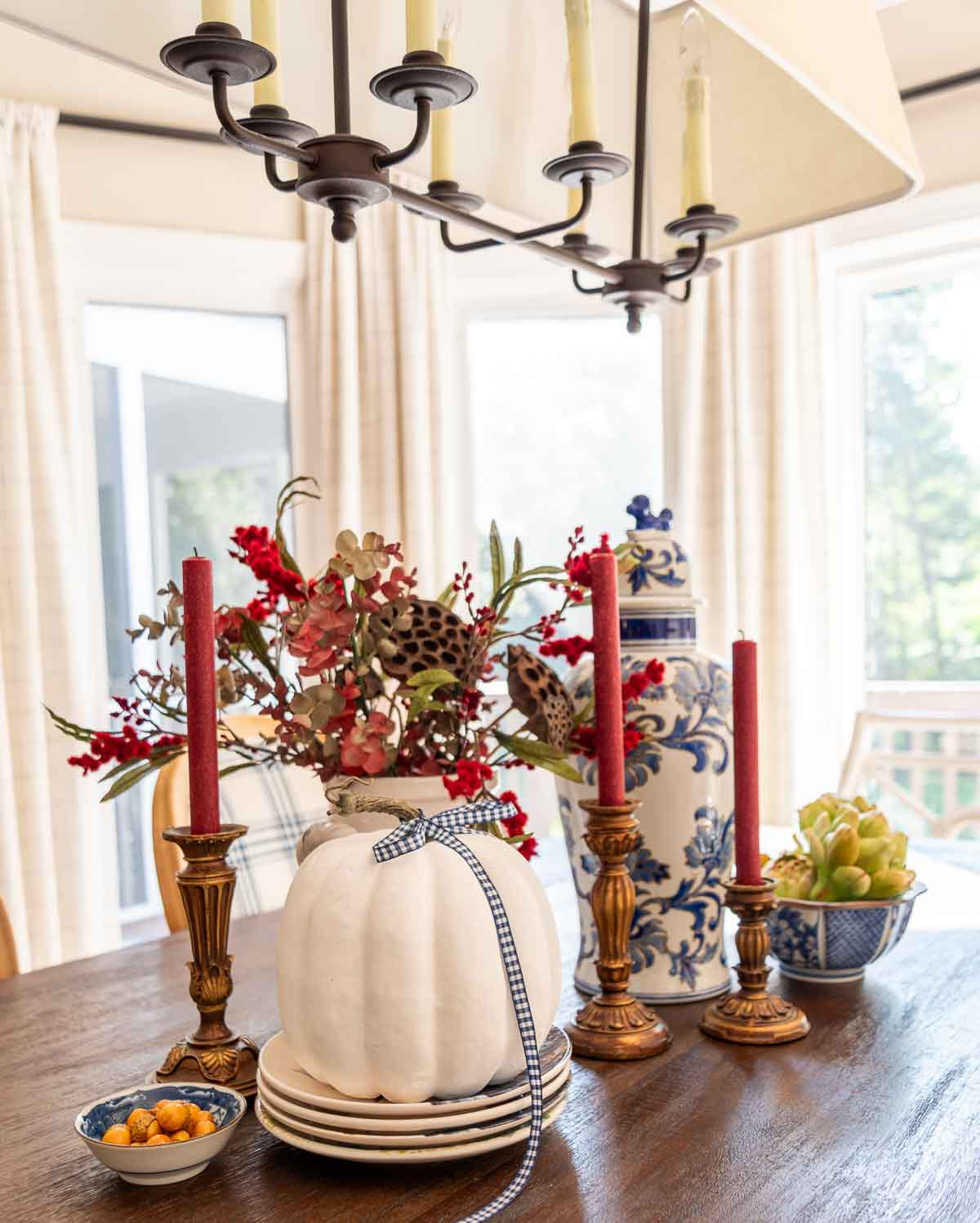 a diy fall table centerpiece using blue, white and burgundy colors, consisting of a large lidded urn, a floral arrangement, and a large white pumpkin on a stack of fall plates