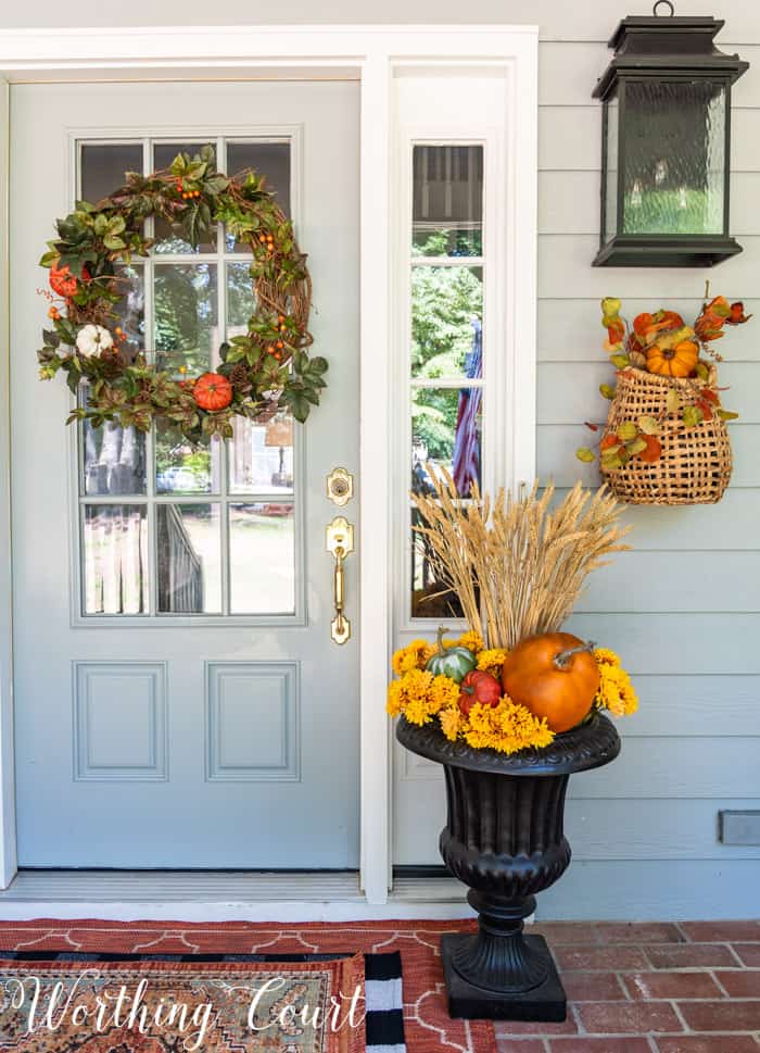 gray front door with a fall wreath flanked by a black urn decorated for fall with pumpkins and wheat stalks