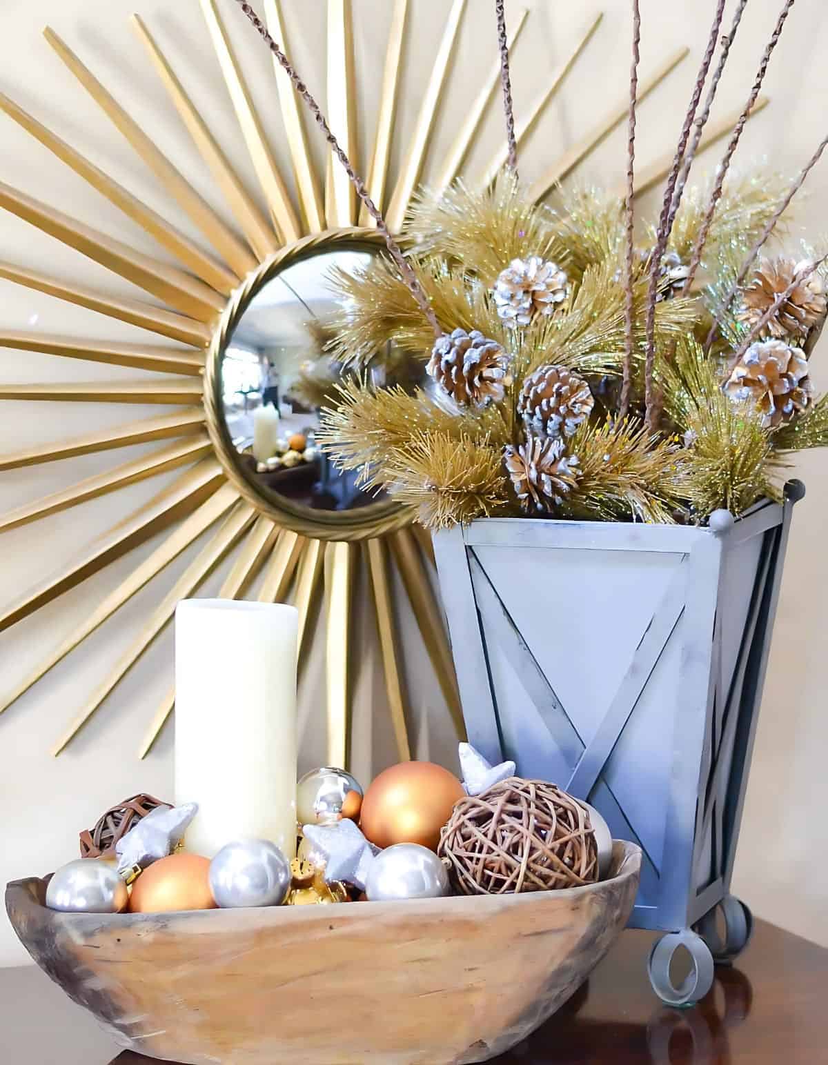 pewter container filled with gold faux pine branches and a small dough bowl filled with gold and silver ornaments in front of a starburst mirror