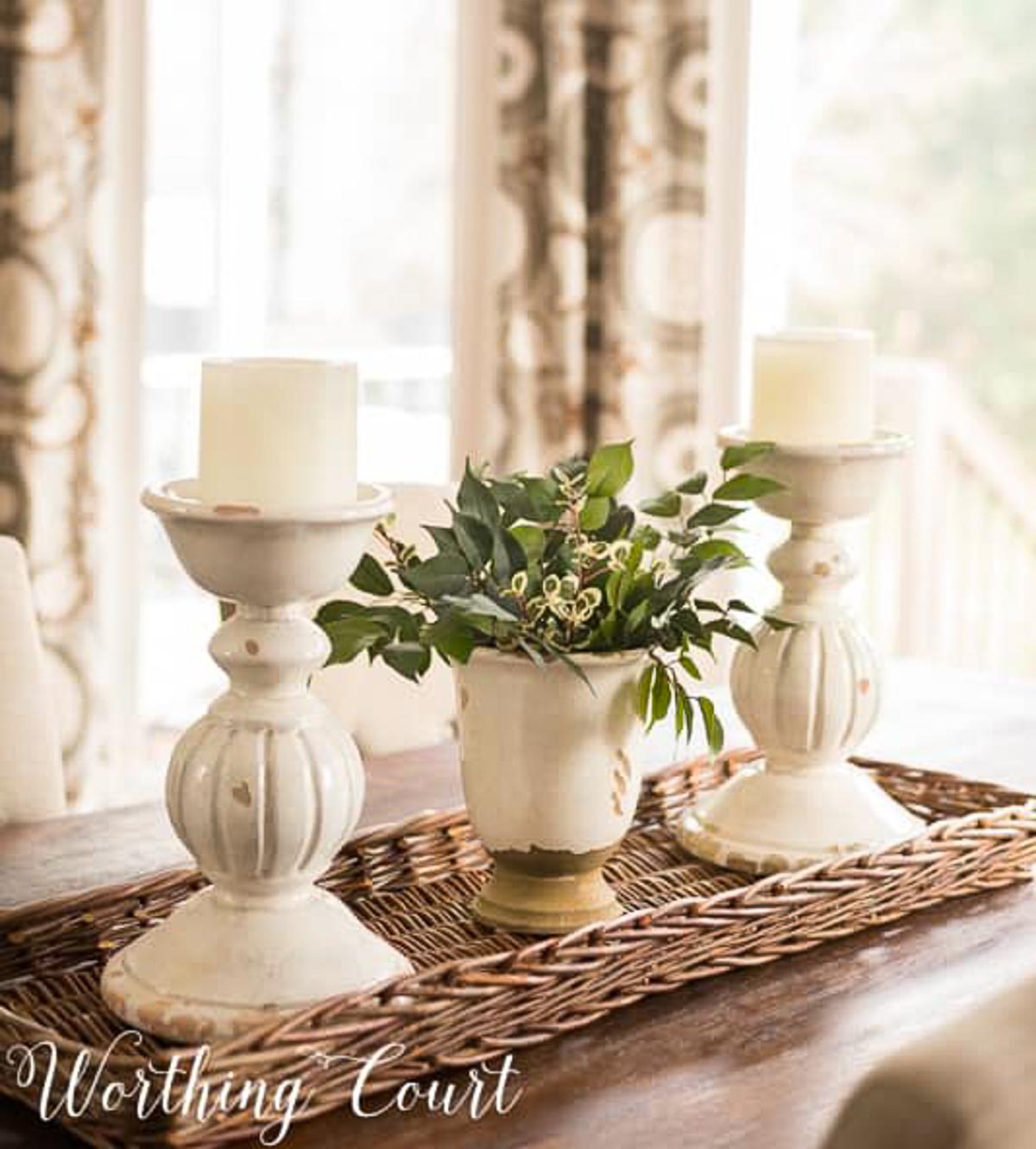 greenery in a white vase between two chunky white candlesticks on a long wicker tray on a dining table