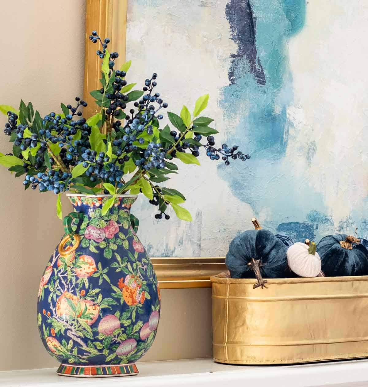 blue floral vase with blue berry stems and a gold planter filled with blue and ivory colored mini pumpkins on a fireplace mantel